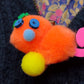 Orange Pom-Pom Gralockamay Pin with hot pink tail, yellow feet,  a light blue nose dark blue eyes and a green flower looking crest.