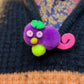 A Purple Gralockamay Pom-Pom pin with a hot pink tail a yellow nose, neon yellow feet, light pink eyes and a green flower hair tuff. The pin in facing forward .