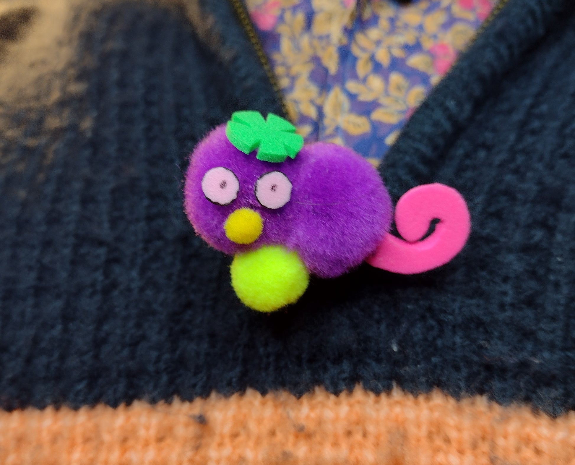 A Purple Gralockamay Pom-Pom pin with a hot pink tail a yellow nose, neon yellow feet, light pink eyes and a green flower hair tuff. The pin in facing forward .